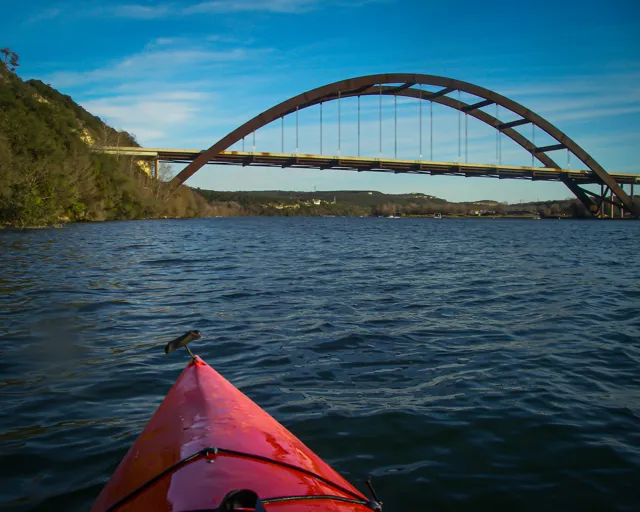 The bow of a kayak points toward Pennybacker Bridge, placing the viewer out on Lake Austin.